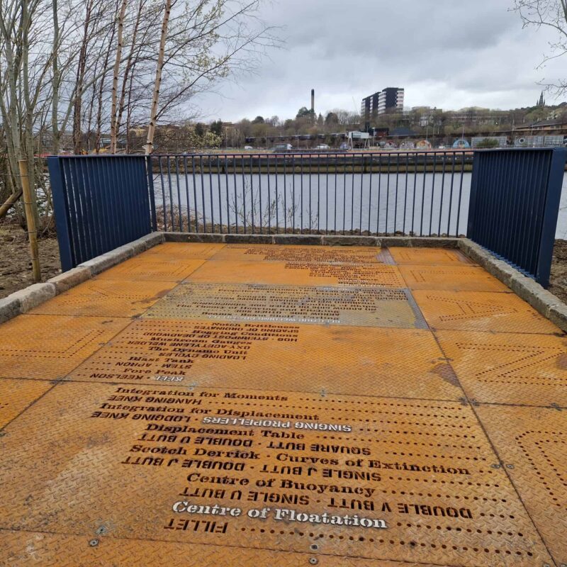 A large slab of ironwork, engraved with writing. A river can be seen in the background.