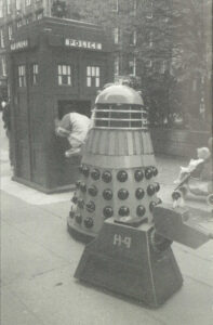 Black and white photo of the Buchanan Street police box with someone peering in the door catch. A Dalek and K9 stand sentry outside.