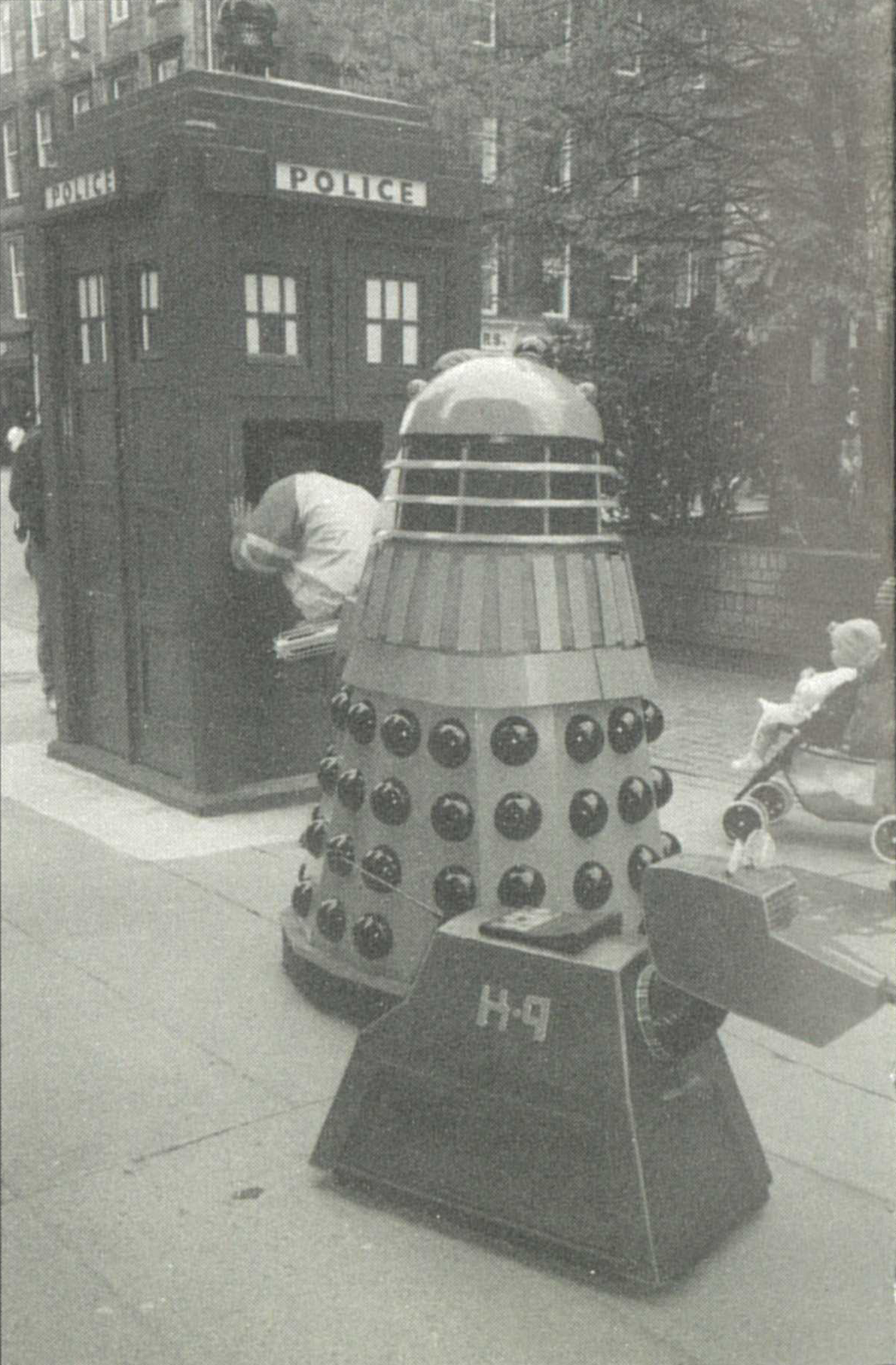 Black and white photo of the Buchanan Street police box with someone peering in the door catch. A Dalek and K9 stand sentry outside.