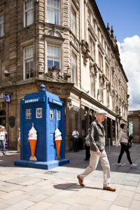 Passers by walk past a blue police box on Buchanan Street on sunny day. The police box is decorated with large ice cream cones on it's sides.