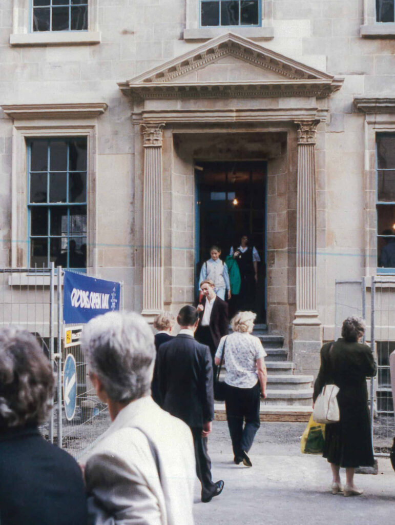 People standing in front of the doorway of a large stone building. A blue sign reading 'Doors Open' is beside the door.